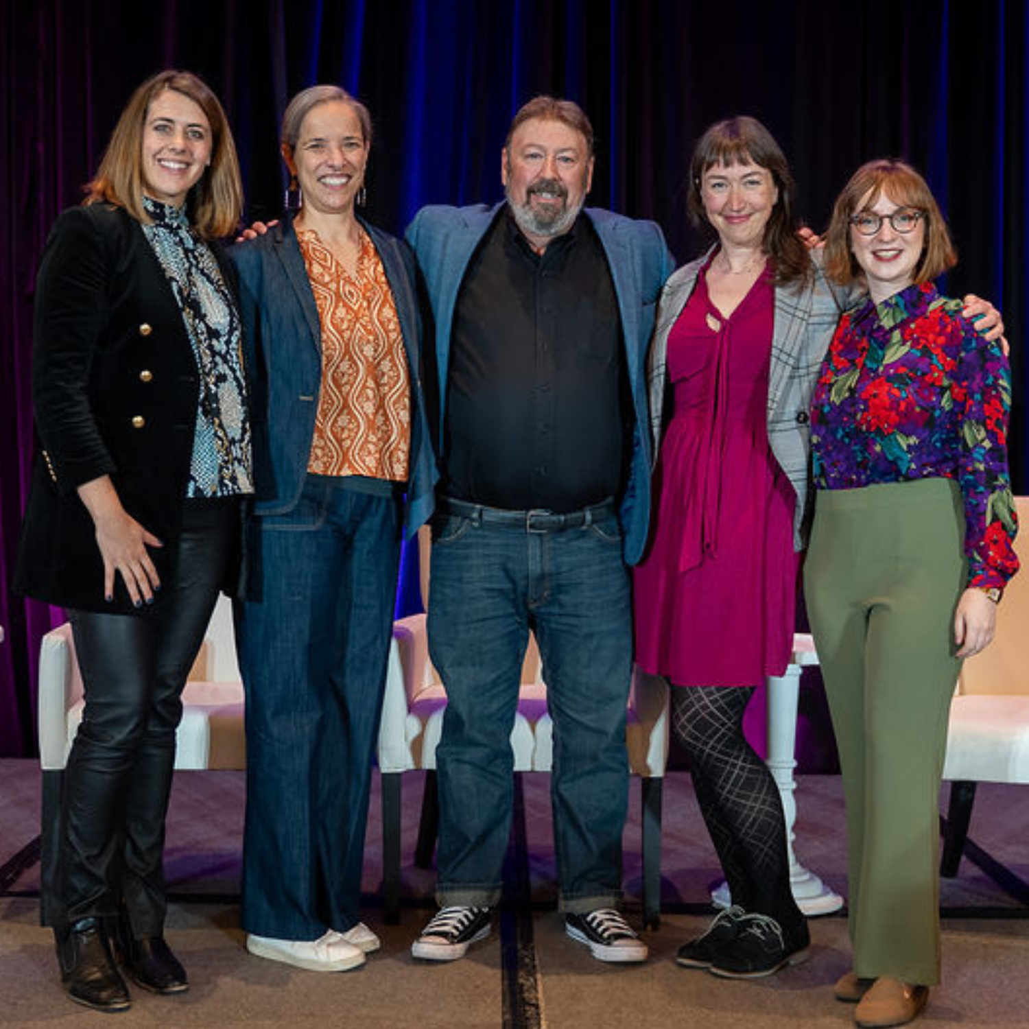 Four white middle aged women with a mille aged white male in the center of a group photograph on stage at a conference.