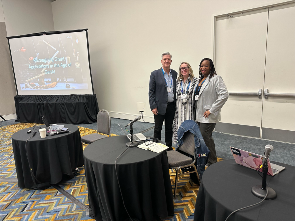 Middle aged white male with short, silver hair stands to the left of a middle aged, white woman with blonde hair and a younger black woman with shoulder length dark dreadlocked hair. They stand next to a presentation screen in front of three tables.