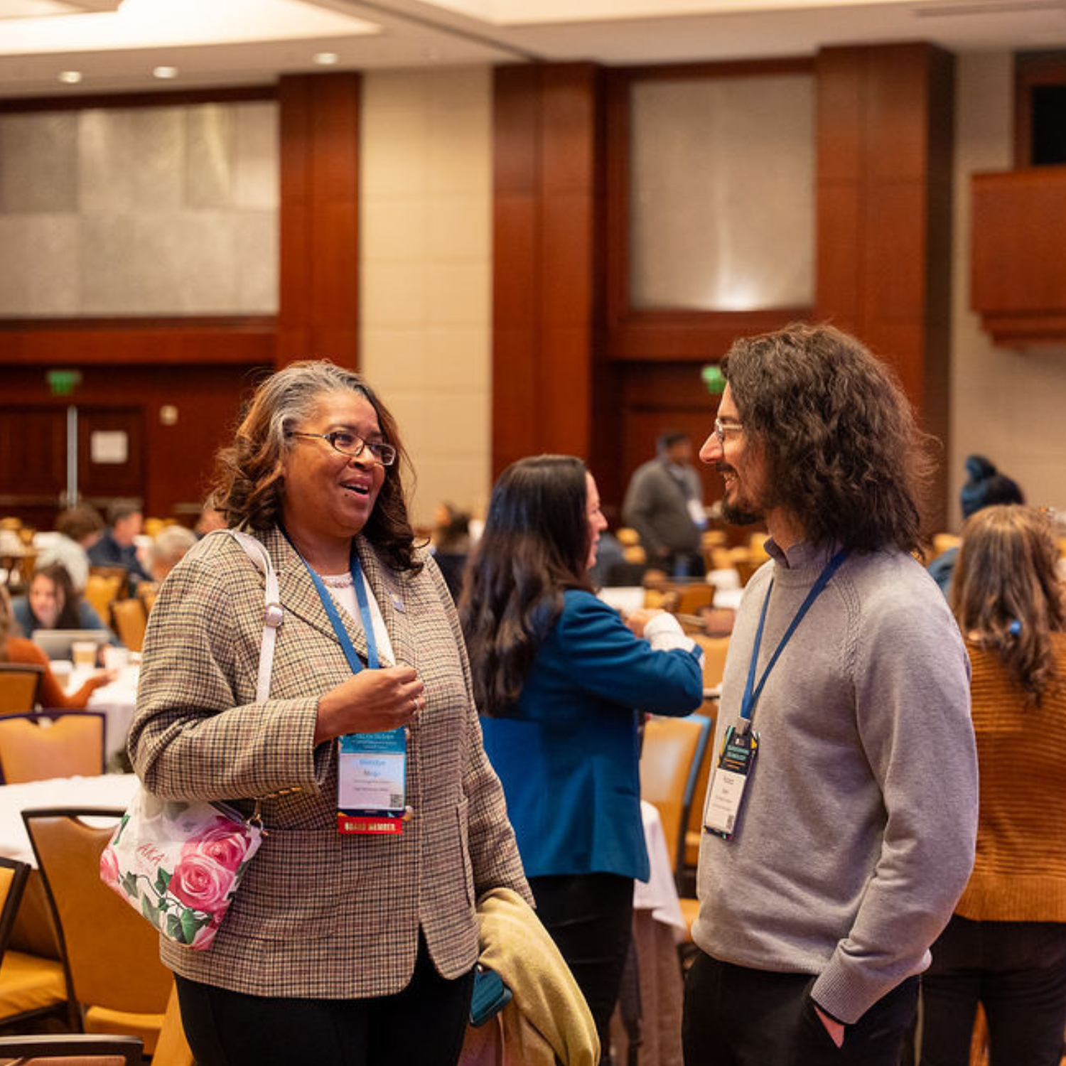 Middle aged black woman with greying hair talking to a younger white male with shoulder length dark hair.