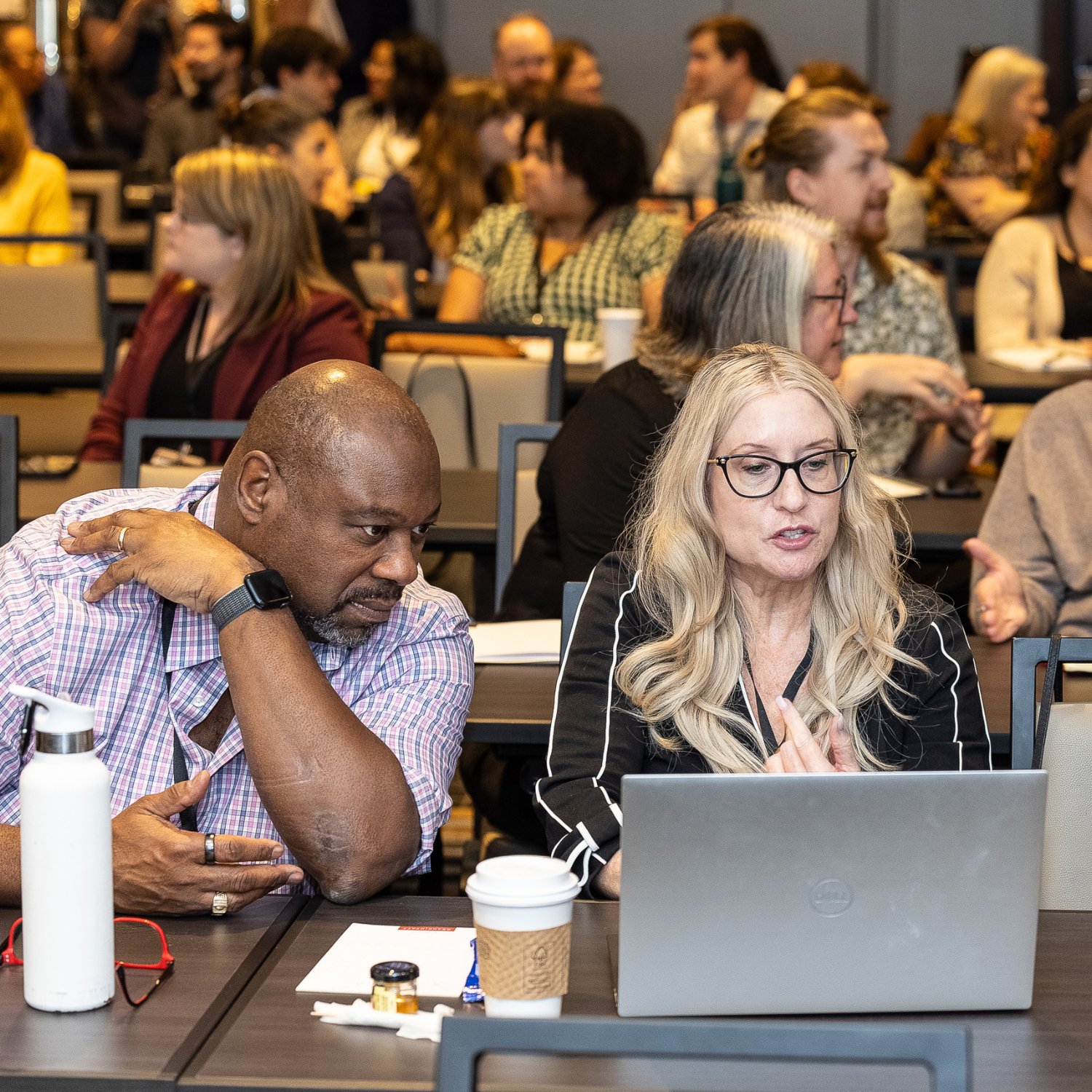 Summit_InfoBlock_Members Middle aged bald black man leans over a laptop to look at the screen as a middle aged white woman with blonde hair speaks.